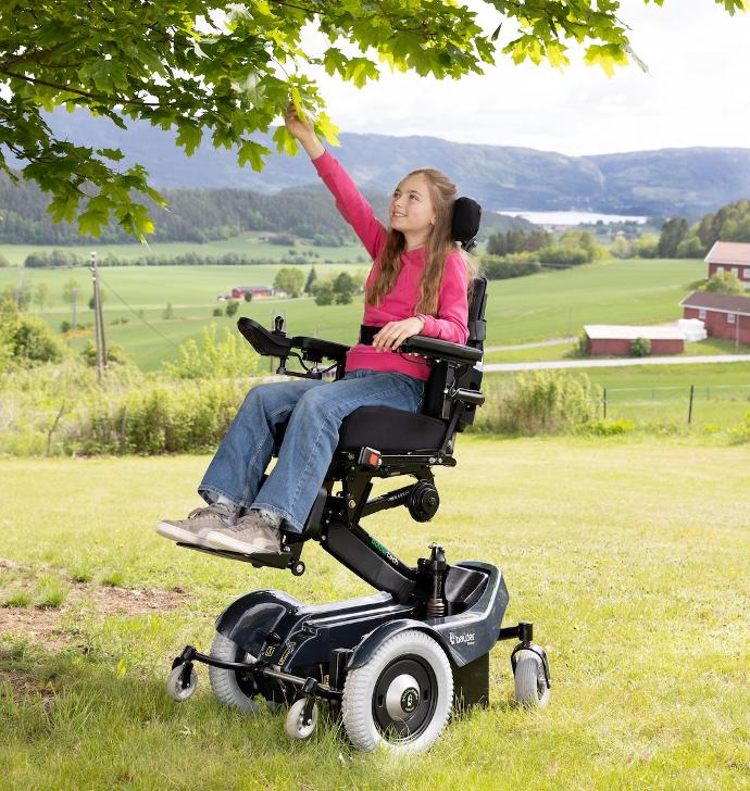 A young girl sitting in a Balder J340 childrens powerchair. She is using the elevation function on the wheelchair and reaching leaves on a tree.