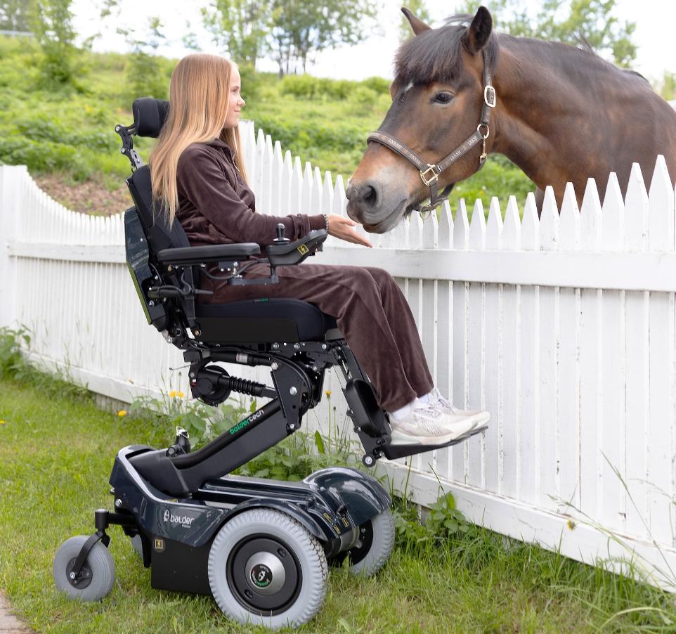 A young girl sitting in a Balder J335 childrens powerchair. She is using the elevation function on the wheelchair and reaching out to a horse.