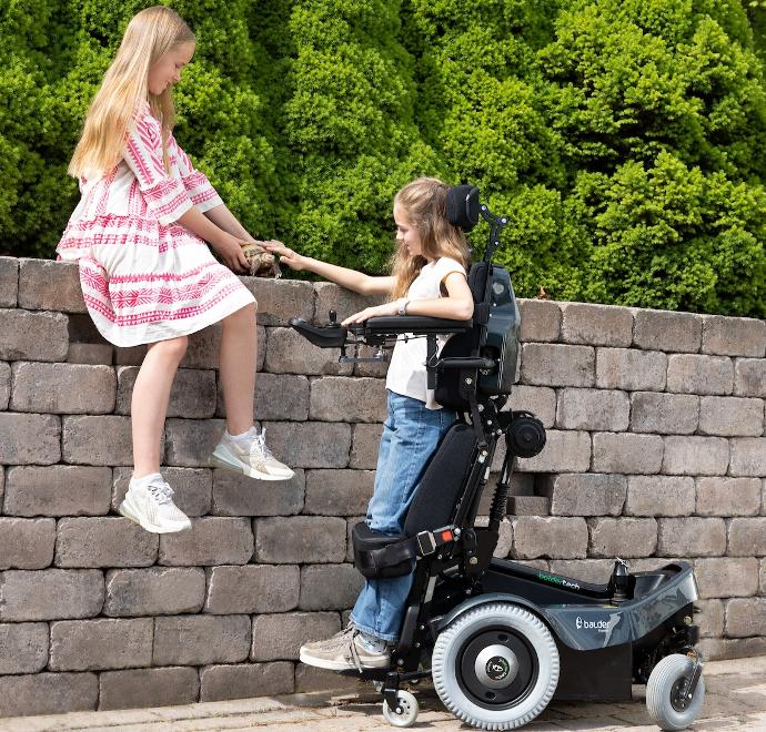 A young girl using the standing function of a Balder Junior J340 childrens standing powerchair. She is with another young girl who is sitting on a high wall. Together they are petting a tortoise.